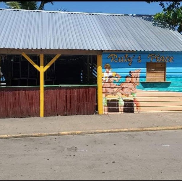 A colorful wooden building on Roatan, Honduras, features a mural of two men sitting at a bar with the sign "Ruty's Place" and an open window, with outdoor seating under a corrugated metal roof.