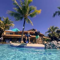 A tropical resort in Roatan, Honduras, features a swimming pool surrounded by palm trees, a wooden deck with outdoor seating, and a spacious building with a thatched roof in the background.