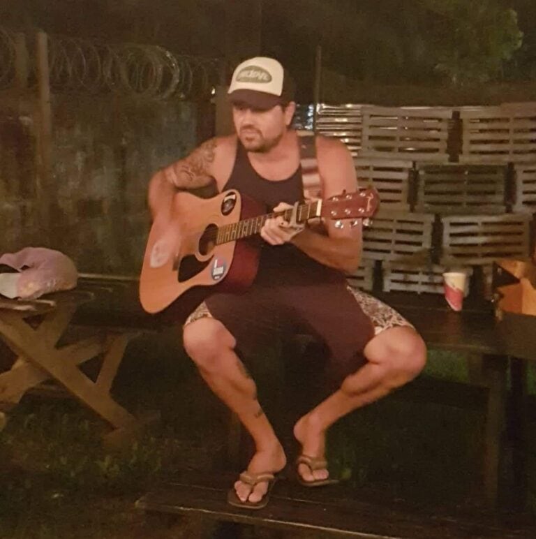A man wearing a cap, tank top, and shorts plays an acoustic guitar in an outdoor setting in Roatan, Honduras, with wooden crates and barbed wire visible in the background.