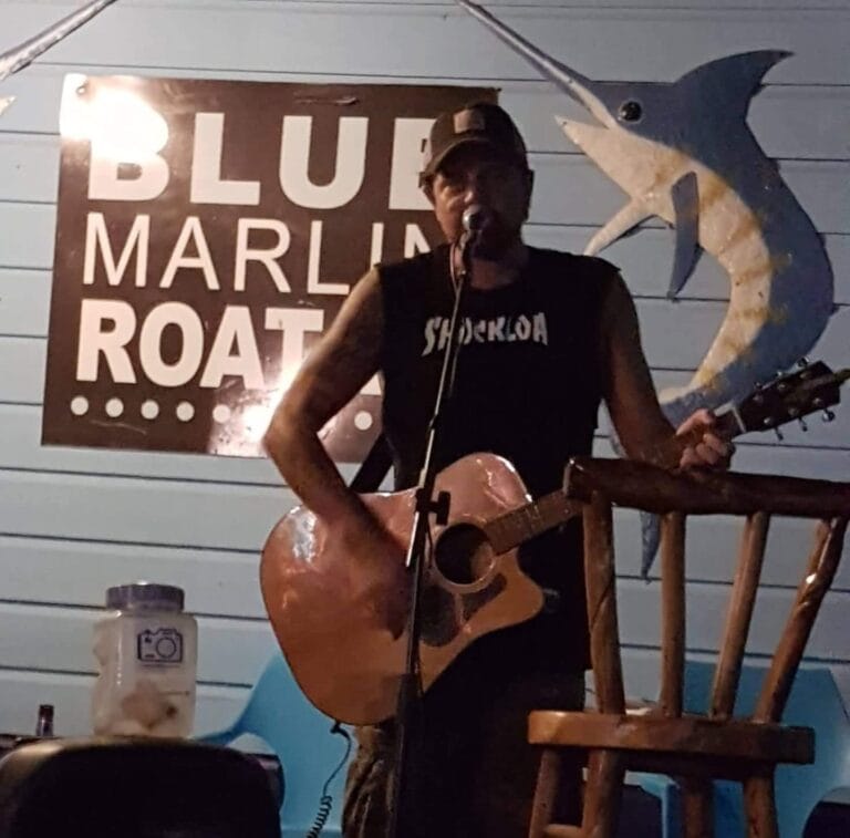A person performing live music with an acoustic guitar at a beachfront bar or restaurant in Roatan, Honduras, with a sign reading "Blue Marlin Roatan" and a decorative marlin fish on the wall behind them.