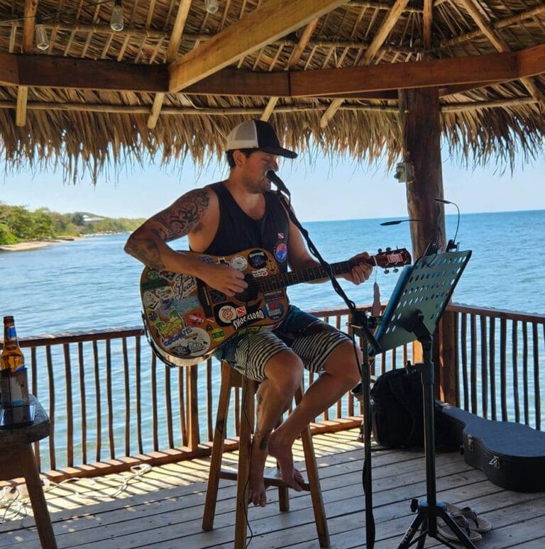 A man with tattoos playing an acoustic guitar decorated with colorful stickers, singing into a microphone under a thatched-roof structure on a wooden deck overlooking the ocean in Roatan, Honduras.
