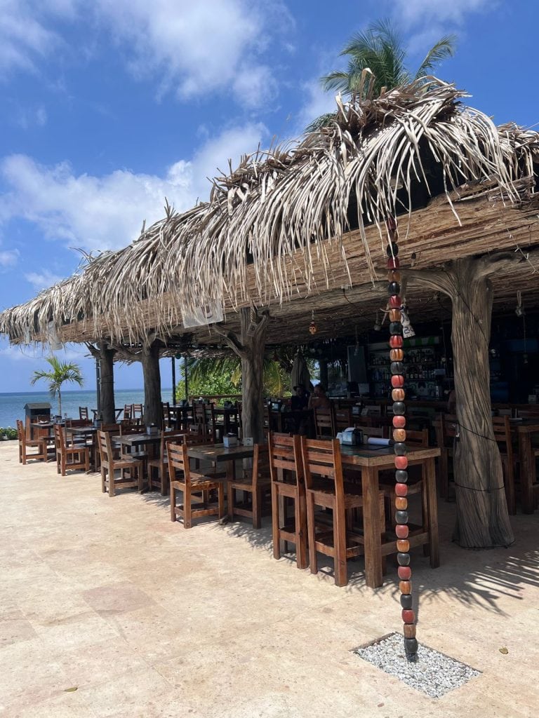 A beachfront open-air restaurant in Roatan, Honduras, features wooden tables and chairs under a thatched palm frond roof with ocean views and a blue sky in the background.
