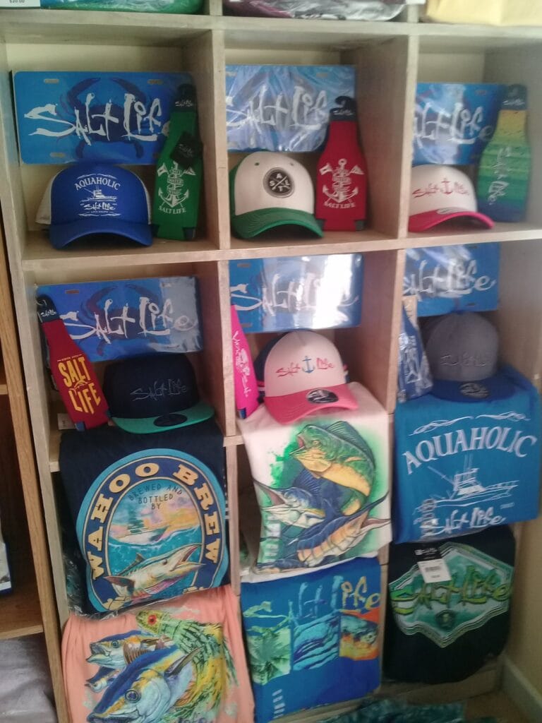 Sailing-themed hats, t-shirts, and artwork featuring marine life are displayed on a wooden shelf at a business in Roatan, Honduras.