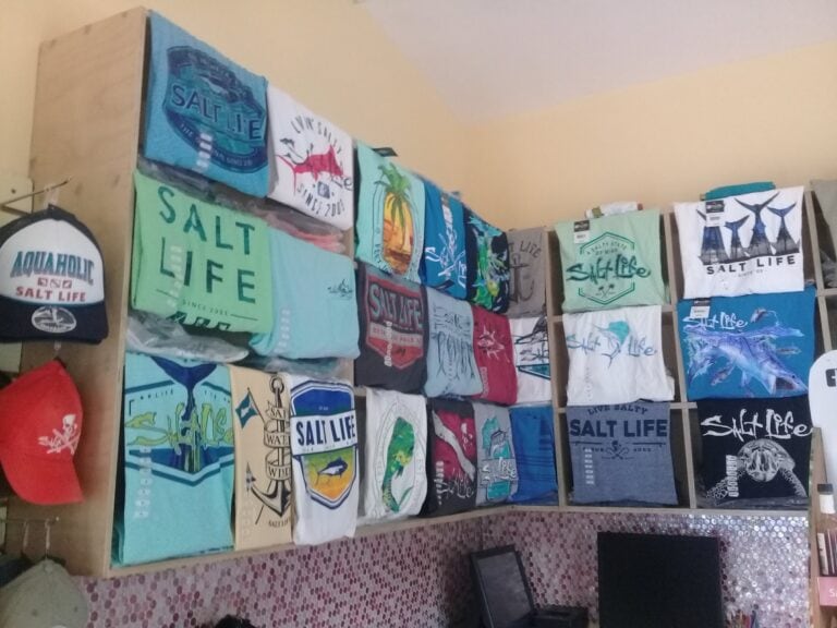 A display of colorful T-shirts and hats featuring the "Salt Life" brand is arranged on a wooden rack inside a shop in Roatan, Honduras.