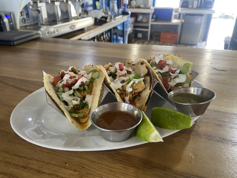 Three tacos filled with meat, lettuce, tomatoes, and cheese are served on a white plate with lime wedges and small metal cups of salsa and green sauce in a restaurant setting in Roatan, Honduras.