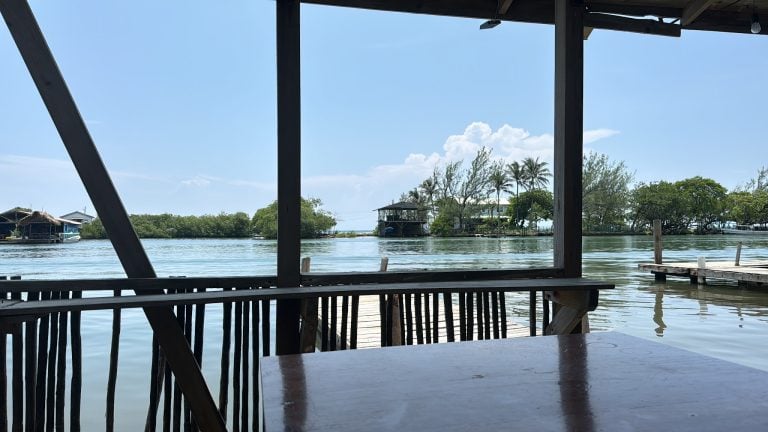 A view of Roatan, Honduras from a wooden dock or boat station overlooking calm water, with houses and trees along the shoreline and a bright blue sky with some clouds in the background.