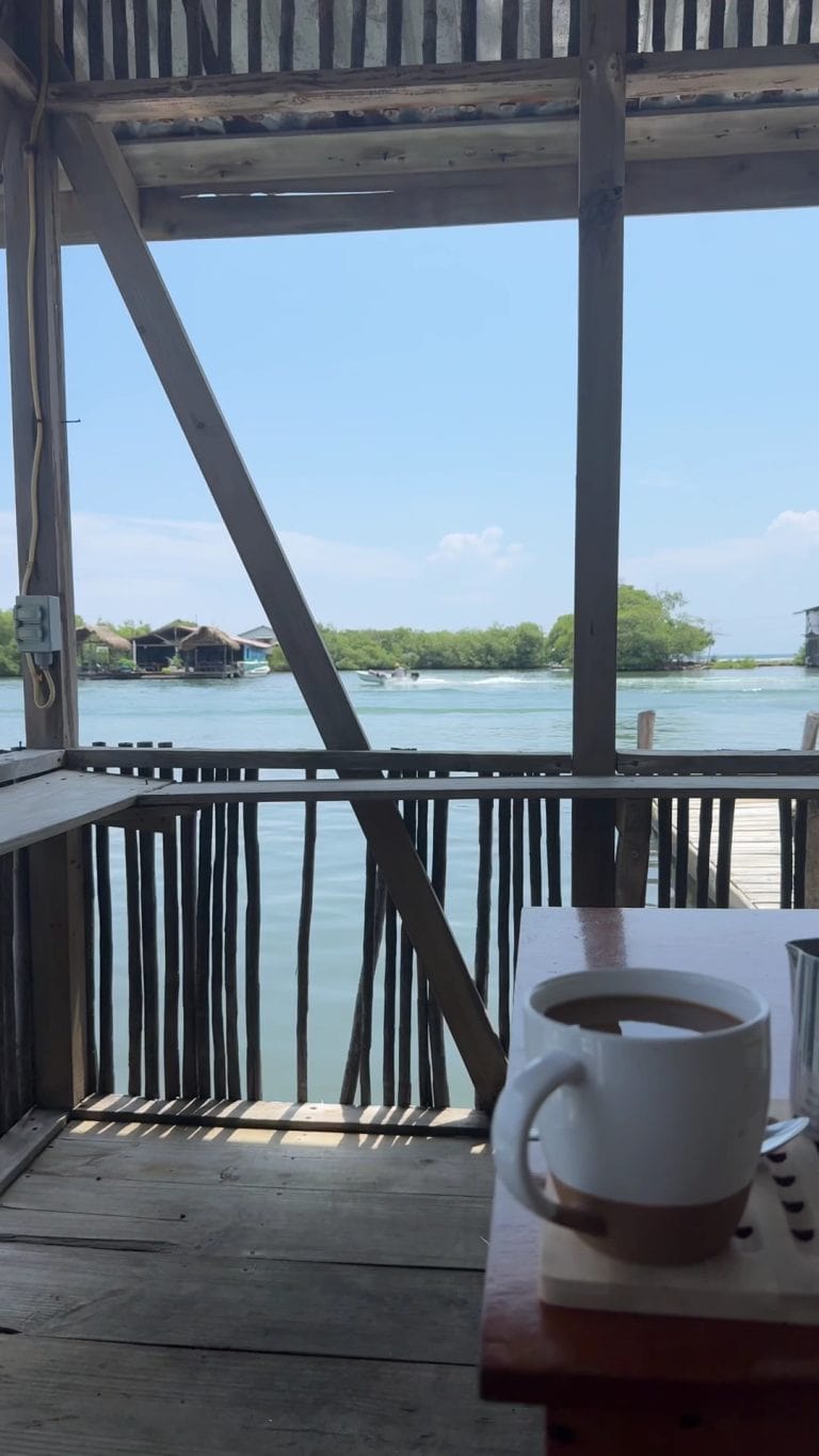 A wooden deck overlooking Roatan's waterfront in Honduras features a white coffee mug on a table in the foreground, with overwater bungalows and boats visible on the calm water under a clear blue sky.