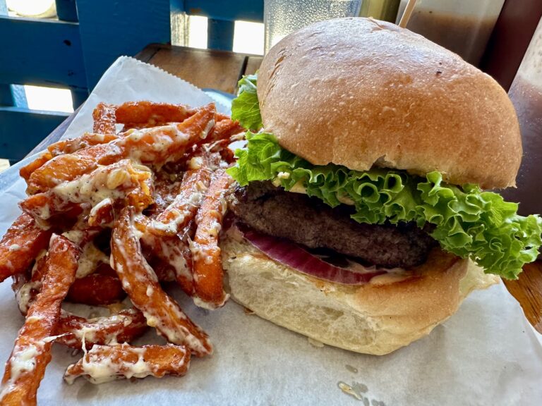 A sandwich with lettuce, tomato, and meat is placed on a bun in front of a window with blue-painted woodwork on Roatan, Honduras.