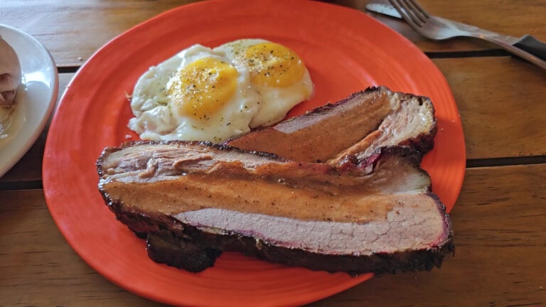A plate of breakfast features two fried eggs seasoned with black pepper and a large, smoked cut of beef ribs served on a vibrant orange plate on a wooden table in Roatan, Honduras.