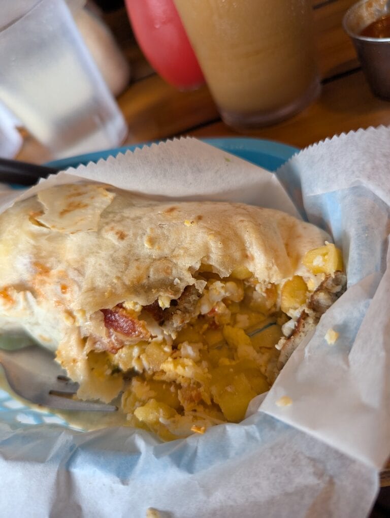 A partially eaten breakfast burrito with eggs, bacon, and potatoes wrapped in foil, set on a table with beverages in the background in Roatan, Honduras.