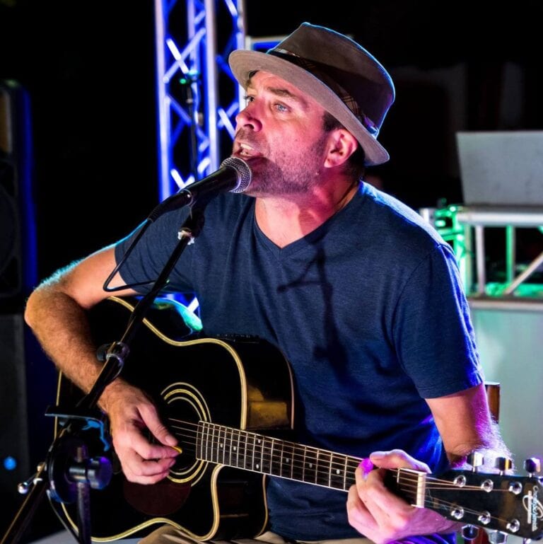 A male musician wearing a fedora hat performs with an acoustic guitar and sings into a microphone on stage with colorful lighting, in Roatan, Honduras.