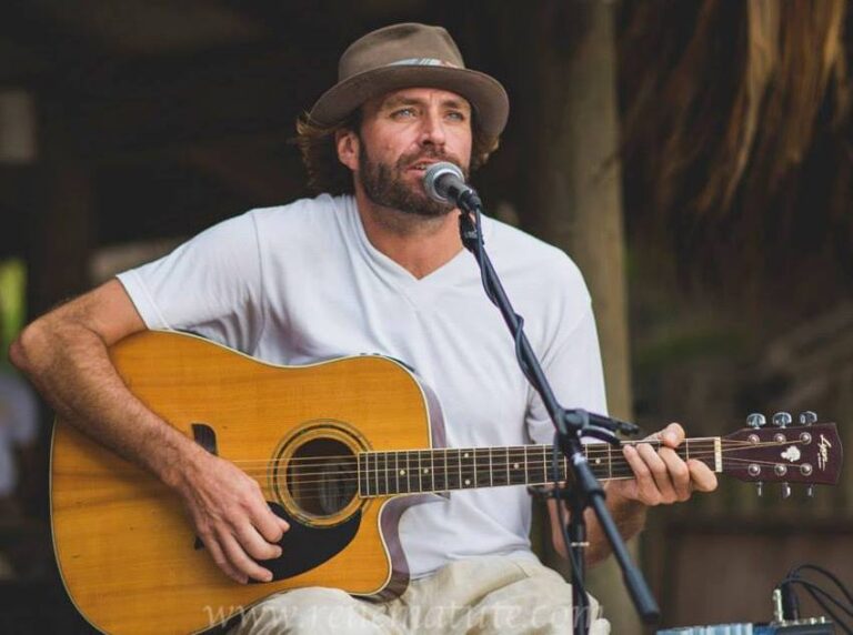 A man wearing a hat and white t-shirt plays an acoustic guitar and sings into a microphone during a musical performance in an outdoor setting in Roatan, Honduras.