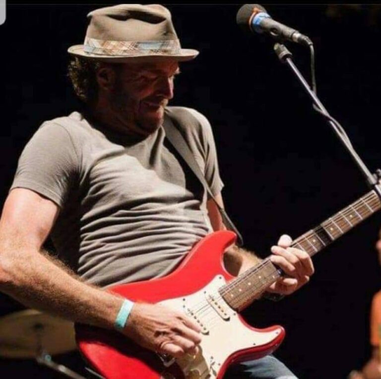 A man wearing a hat and a gray t-shirt plays a red electric guitar on stage in Roatan, Honduras, with a microphone stand nearby and a dark background behind him.
