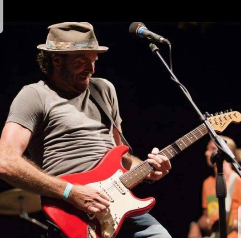 A man wearing a hat and gray shirt is playing a red electric guitar on stage in Roatan, Honduras, with a microphone stand nearby and a blurred person in the background.