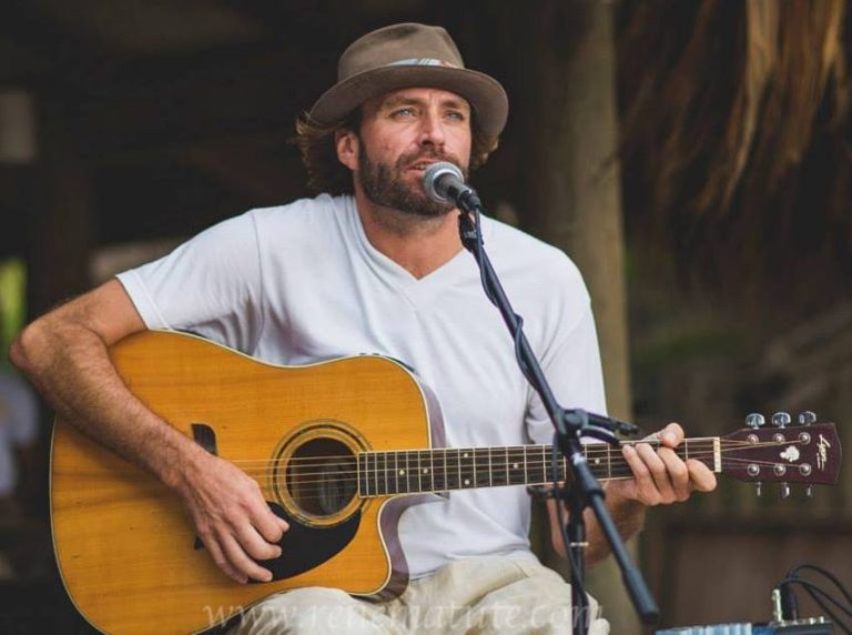A man wearing a brown hat and white t-shirt is performing with an acoustic guitar and singing into a microphone at an outdoor setting in Roatan, Honduras.