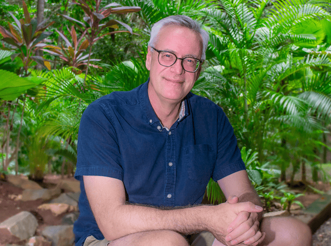 A middle-aged man with glasses and gray hair sitting outdoors among lush tropical plants in Roatan, Honduras.