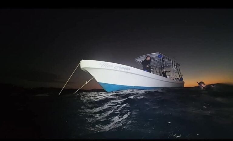 A boat named "Miss Amelia" is anchored in the dark waters near Roatan, Honduras, with two people onboard and one in the water during sunset or nighttime.