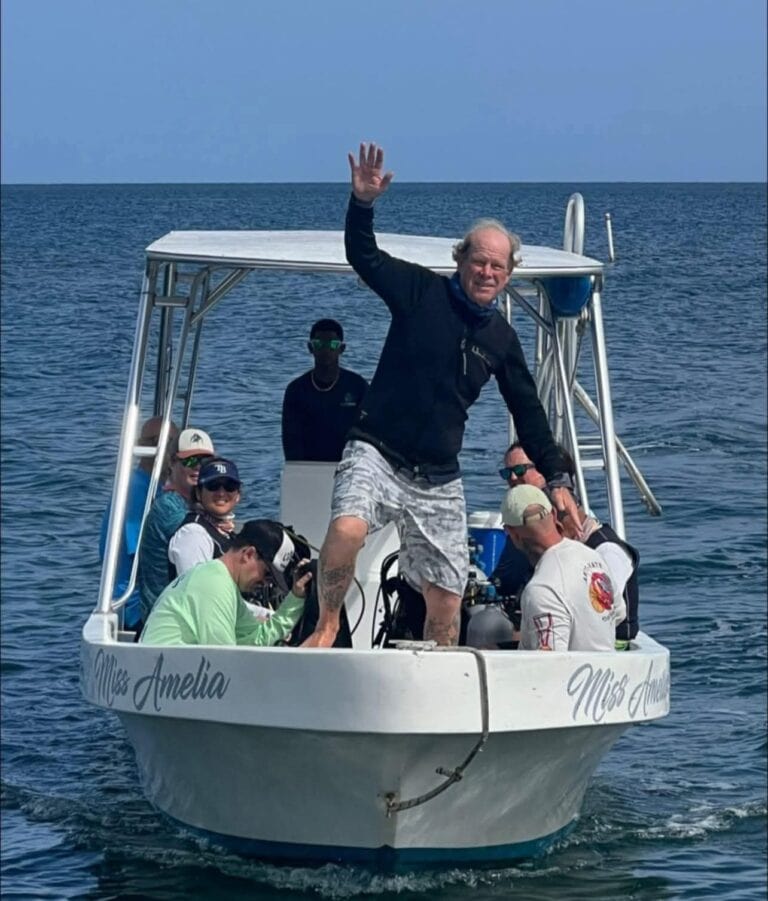 A group of people, including a man waving, enjoying a boat ride labeled "Mrs. Amelia" on the clear blue waters of Roatan, Honduras.