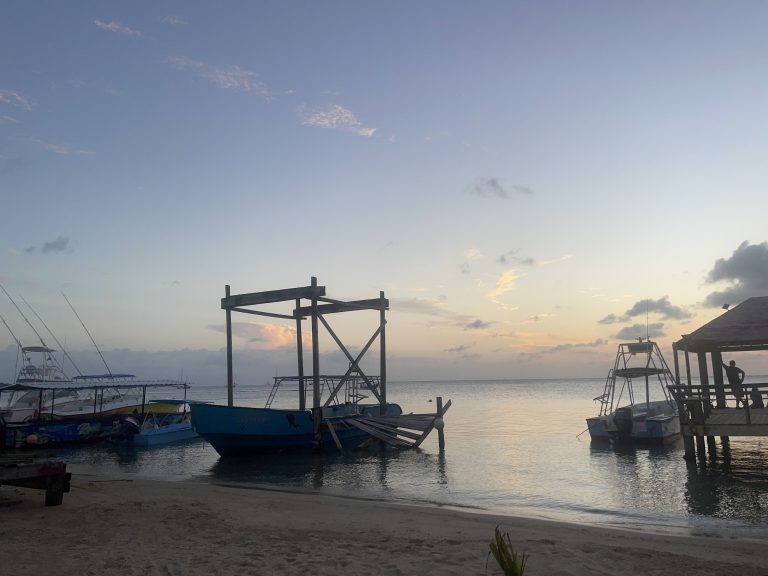 Boats docked at a tranquil harbor in Roatan, Honduras, during sunset with a clear sky and silhouetted structures along the shoreline.