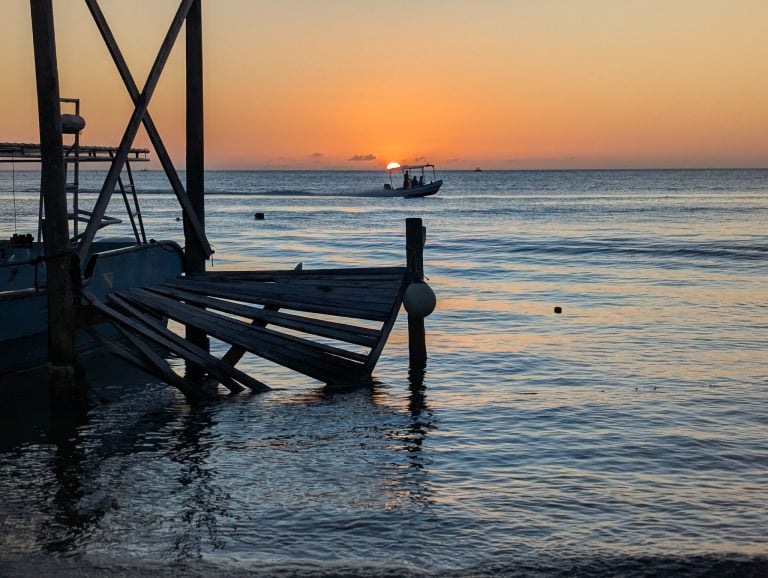 A boat with passengers sailing at sunset on the Caribbean Sea near Roatan, Honduras, with calm water, a dock in the foreground, and a vibrant sky.