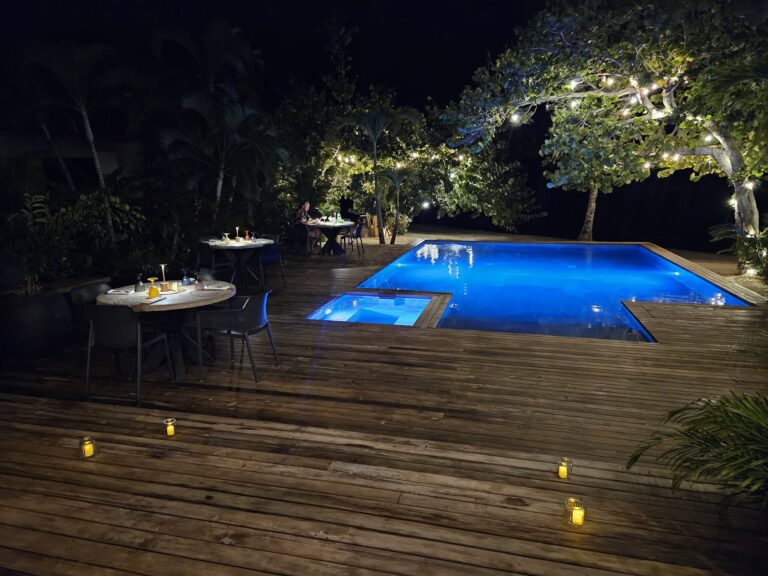 An outdoor dining area on Roatan in Honduras features tables set with candlelit arrangements, a swimming pool illuminated with blue lighting, and lush trees decorated with string lights at night.
