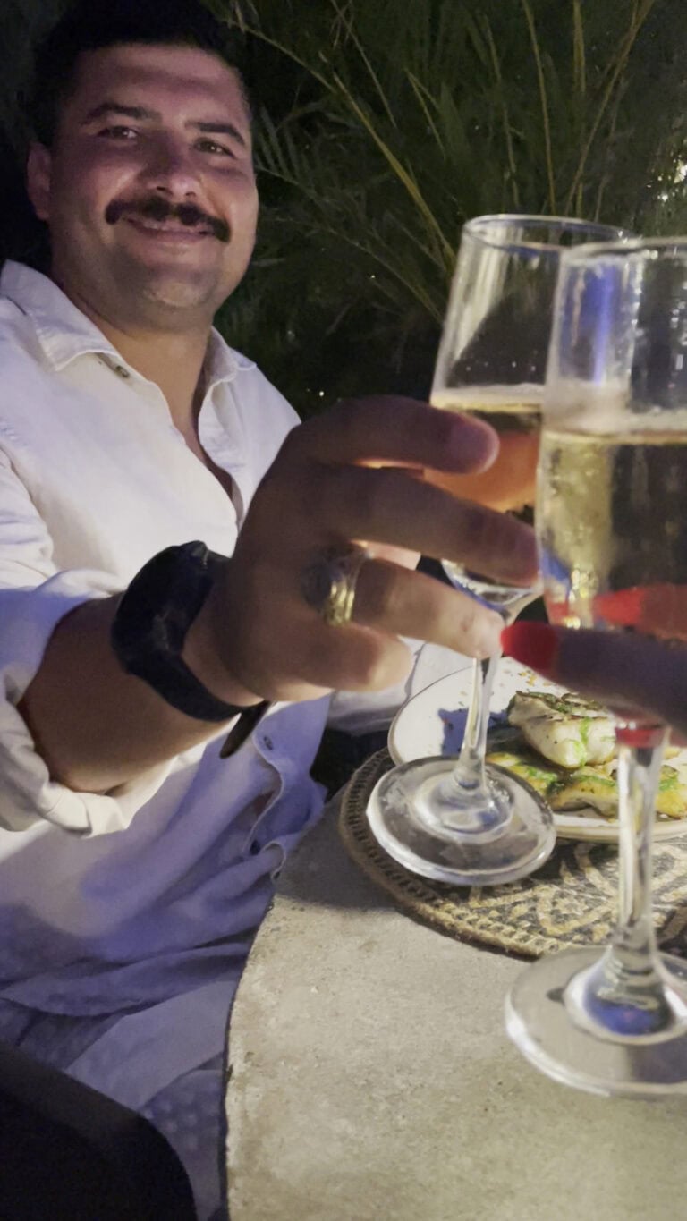 A man in a white shirt and black watch raises a glass of champagne during an evening meal at a restaurant in Roatan, Honduras, with palm foliage in the background and a plate of food on the table.