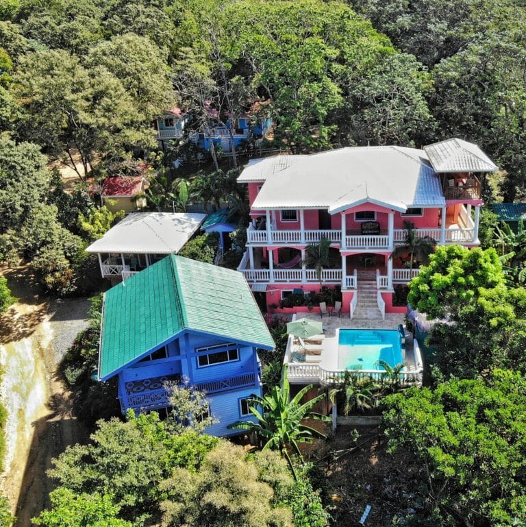 Colorful, two-story houses with white railings and pink, blue, and green exteriors are nestled among lush green trees on Roatan, Honduras.