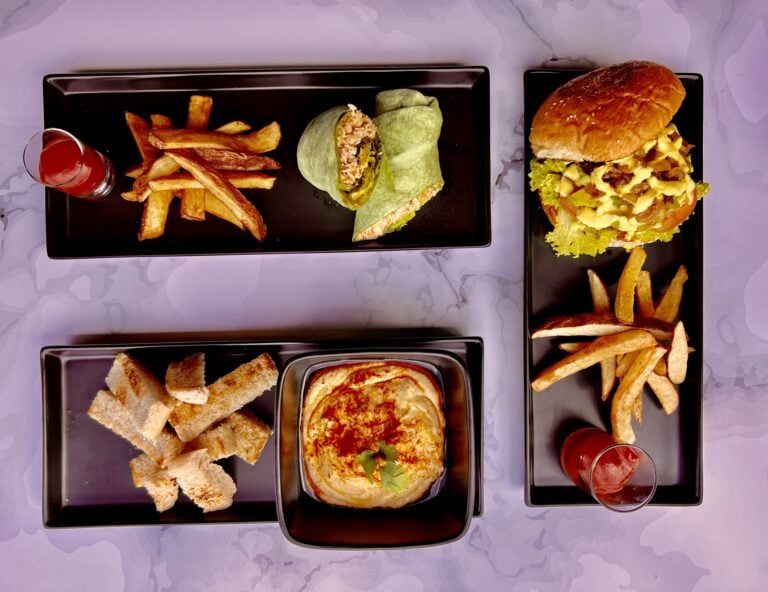 A variety of Mexican-inspired dishes, including fries, a wrap, a burger, and pizza, are served on black trays with small glasses of red drinks on a white tablecloth in Roatan, Honduras.