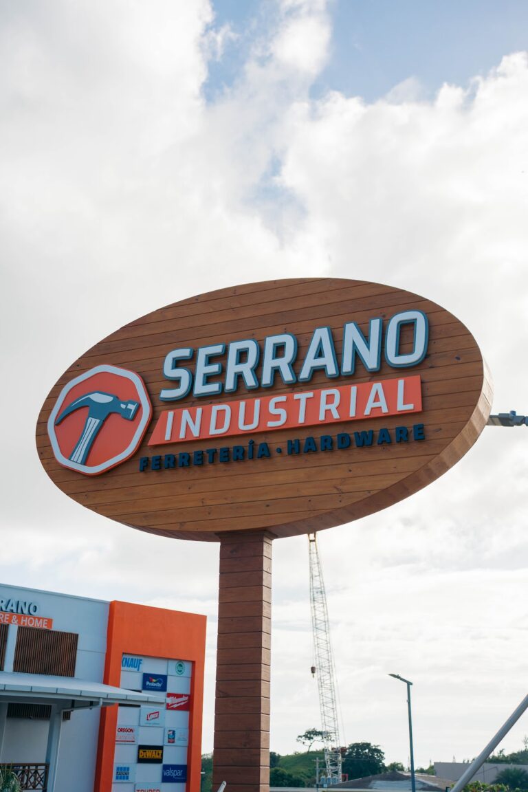 A large wooden sign with blue and red lettering reading "Serrano Industrial" advertises hardware and ferretería services in Roatan, Honduras, with a partly cloudy sky in the background.