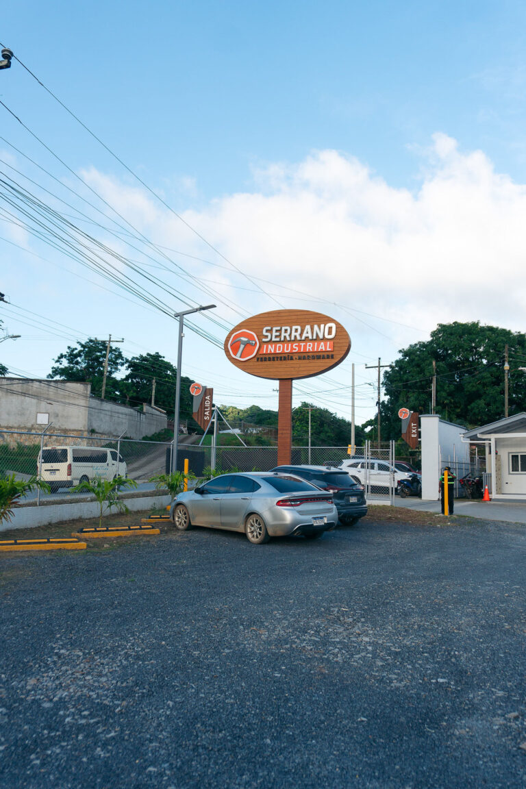 A parking lot in Roatan, Honduras, features several parked cars, a large orange and white Serrano Industrial sign for hardware and DIY supplies, and a metal fence with trees and utility poles in the background under a partly cloudy sky.