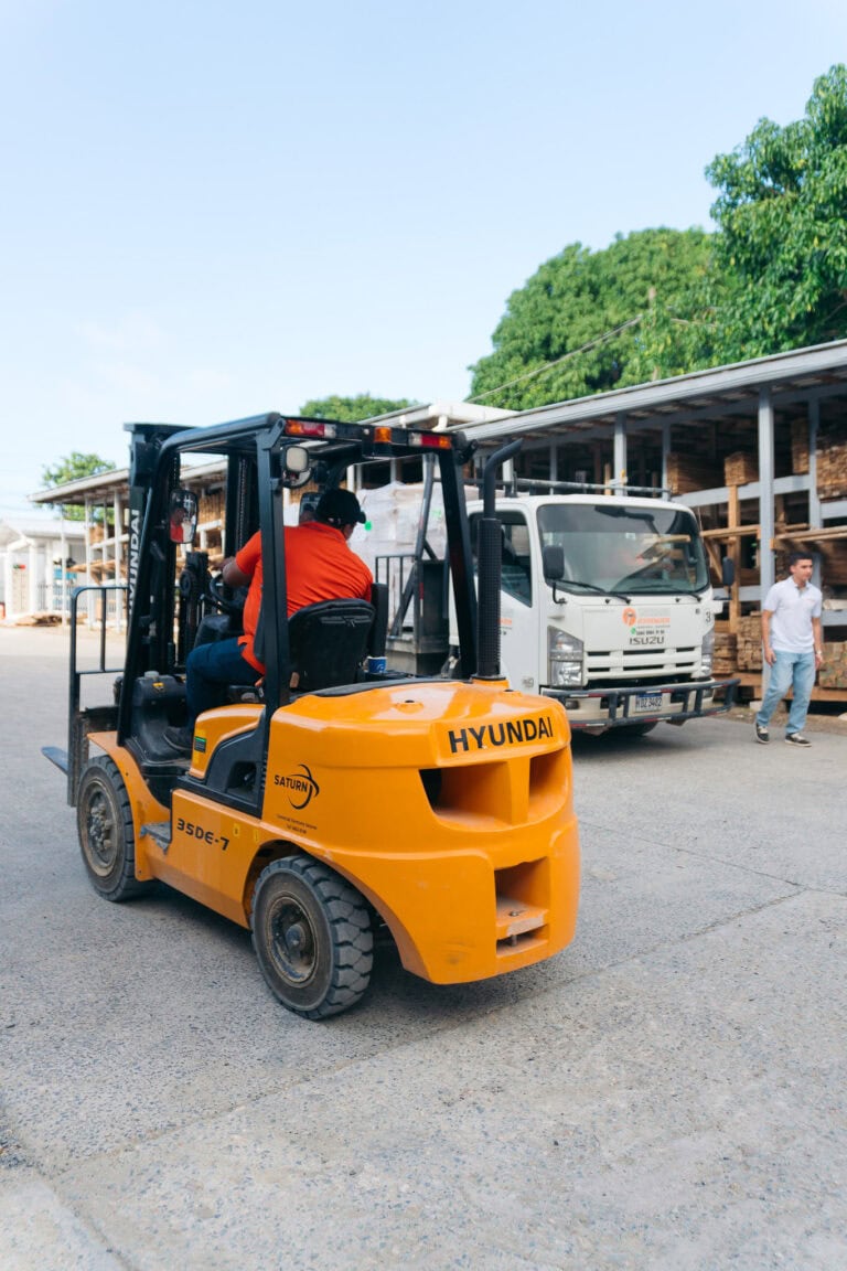 A worker operates a yellow Hyundai forklift transporting cargo in front of a white Isuzu truck at a construction site in Roatan, Honduras, with a partially built wooden structure and lush green trees in the background.