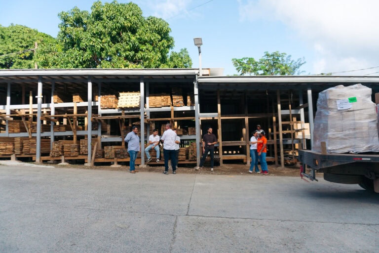 A group of people stands in front of a partially constructed building with stacks of wood on Roatan, Honduras, under a clear blue sky with lush green trees in the background.