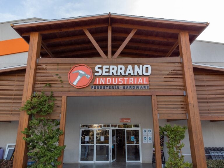 The entrance of Serrano Industrial hardware store in Roatan, Honduras, features a wooden archway with a sign displaying a hammer logo and the store's name, set against a light-colored building with some greenery around.