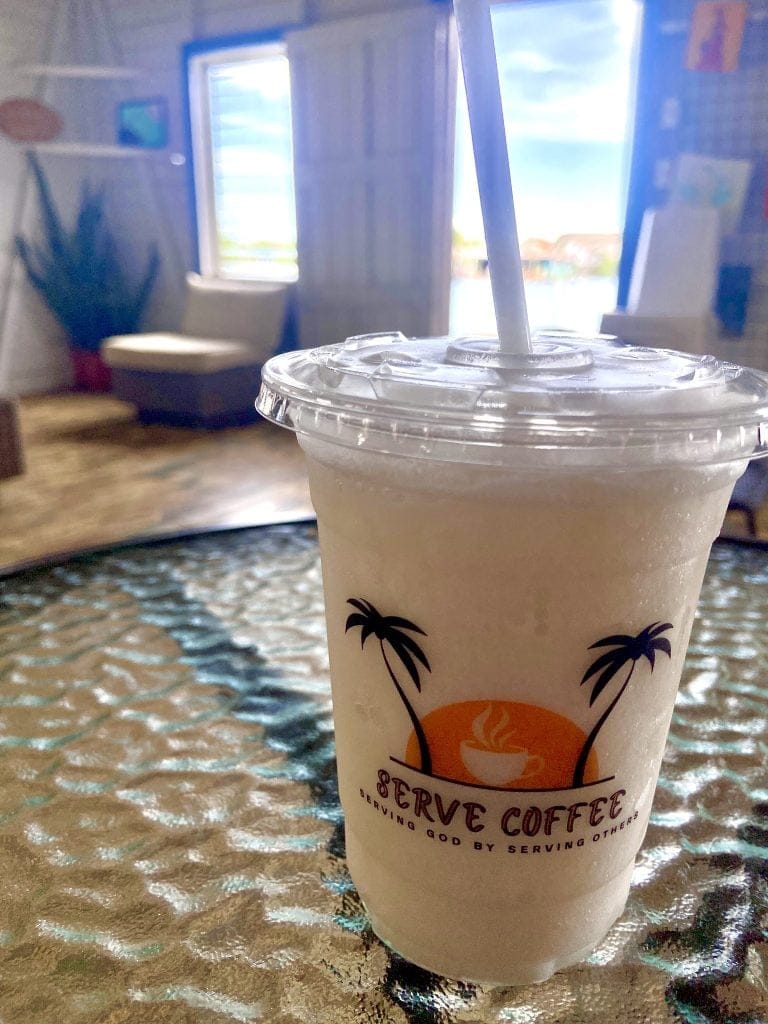 A close-up of an iced coffee in a plastic cup with a straw, featuring a "Roatan Service Coffee" logo, set on a textured glass table inside a casual shop or cafe on Roatan, Honduras, with blurred background windows, posters, and a seating area.