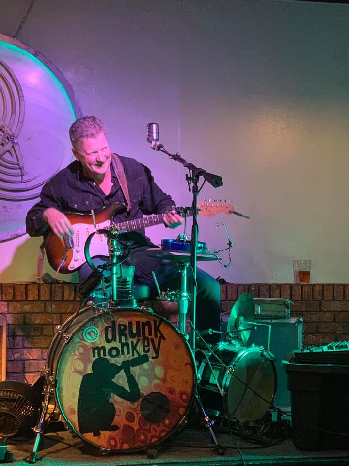 A musician performs live on a drummer set labeled "drunk monkey" with a guitar and microphone on a small stage in Roatan, Honduras, with colorful lighting and a brick wall background.