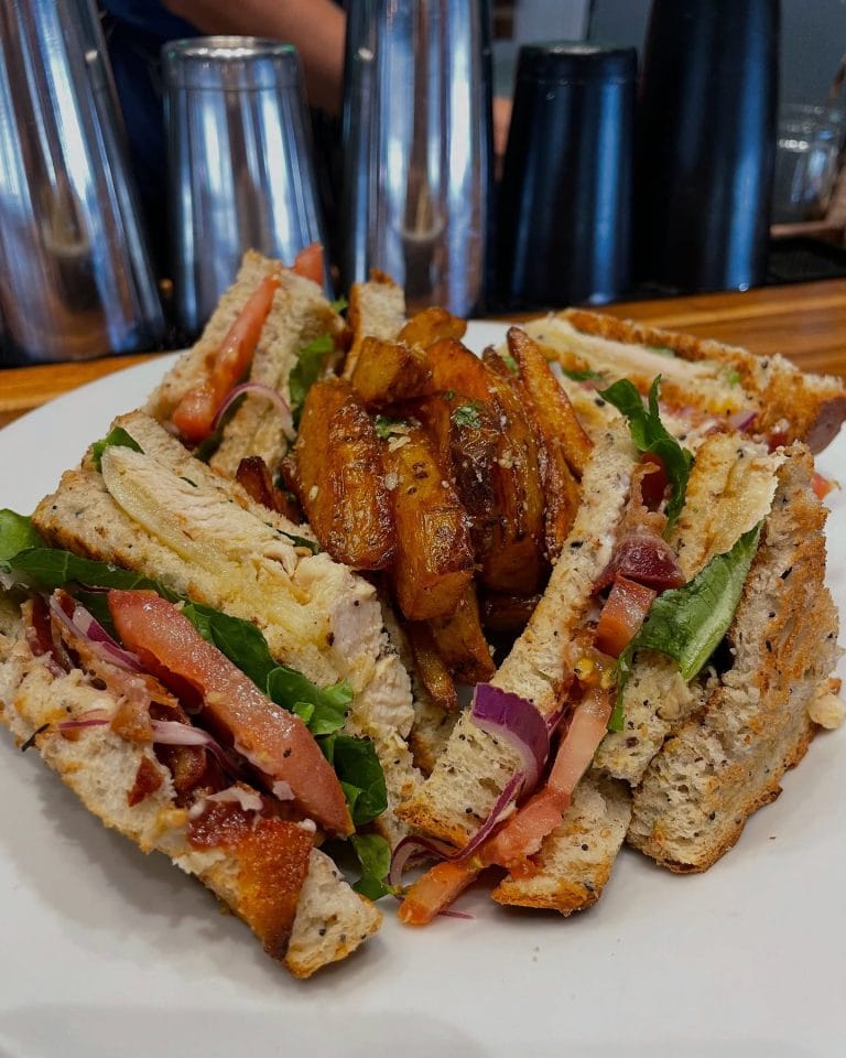 A plate of club sandwich with grilled chicken, fresh vegetables, and crispy fries is shown on a wooden table at a restaurant in Roatan, Honduras.