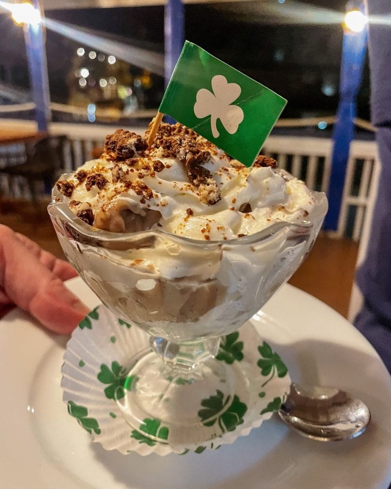 A dessert in a glass bowl garnished with crushed cookies and whipped cream, featuring a small Irish flag, is displayed against a blurred background of a restaurant or bar in Roatan, Honduras.