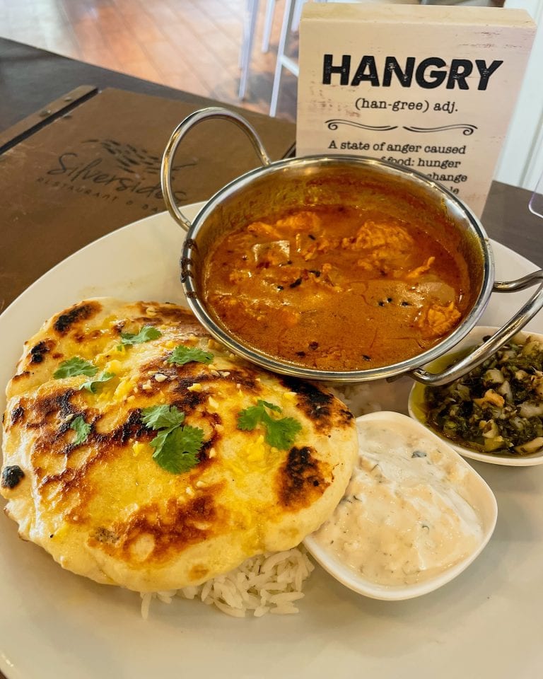 A plate of traditional Honduran cuisine on a table in Roatan, Honduras includes rice, a baked cheese-filled tortilla, a bowl of red meat and vegetable stew, a small dish of creamy sauce, and a side of chopped greens, with a restaurant menu in the background.