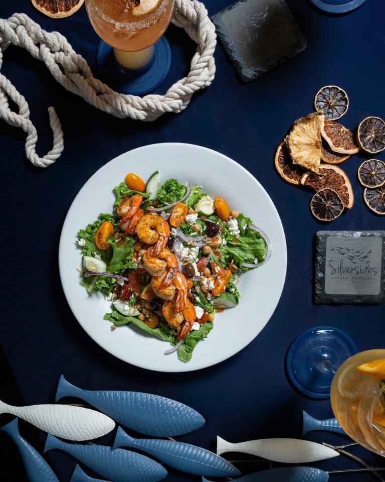 A colorful shrimp and vegetable salad topped with feta cheese and cherry tomatoes is served on a white plate on a dark table at a restaurant in Roatan, Honduras, with dried citrus slices, a cocktail, and decorative rope accents nearby.