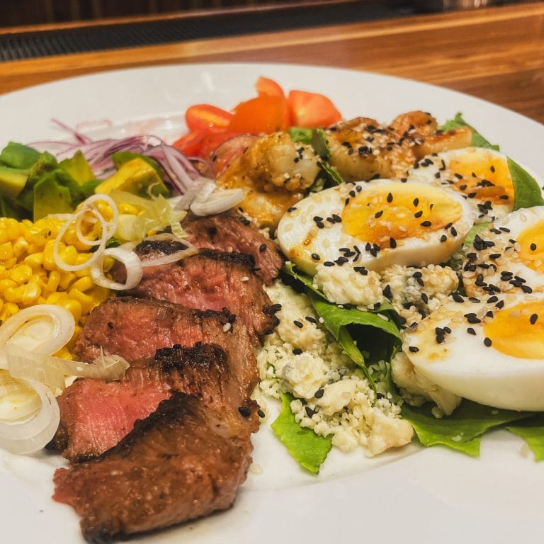 A plate of food featuring sliced medium-rare steak, hard-boiled eggs sprinkled with black sesame seeds, fresh lettuce, chopped tomatoes, thinly sliced onions, corn, and a salad with chopped green peppers, set on a wooden table in Roatan, Honduras.