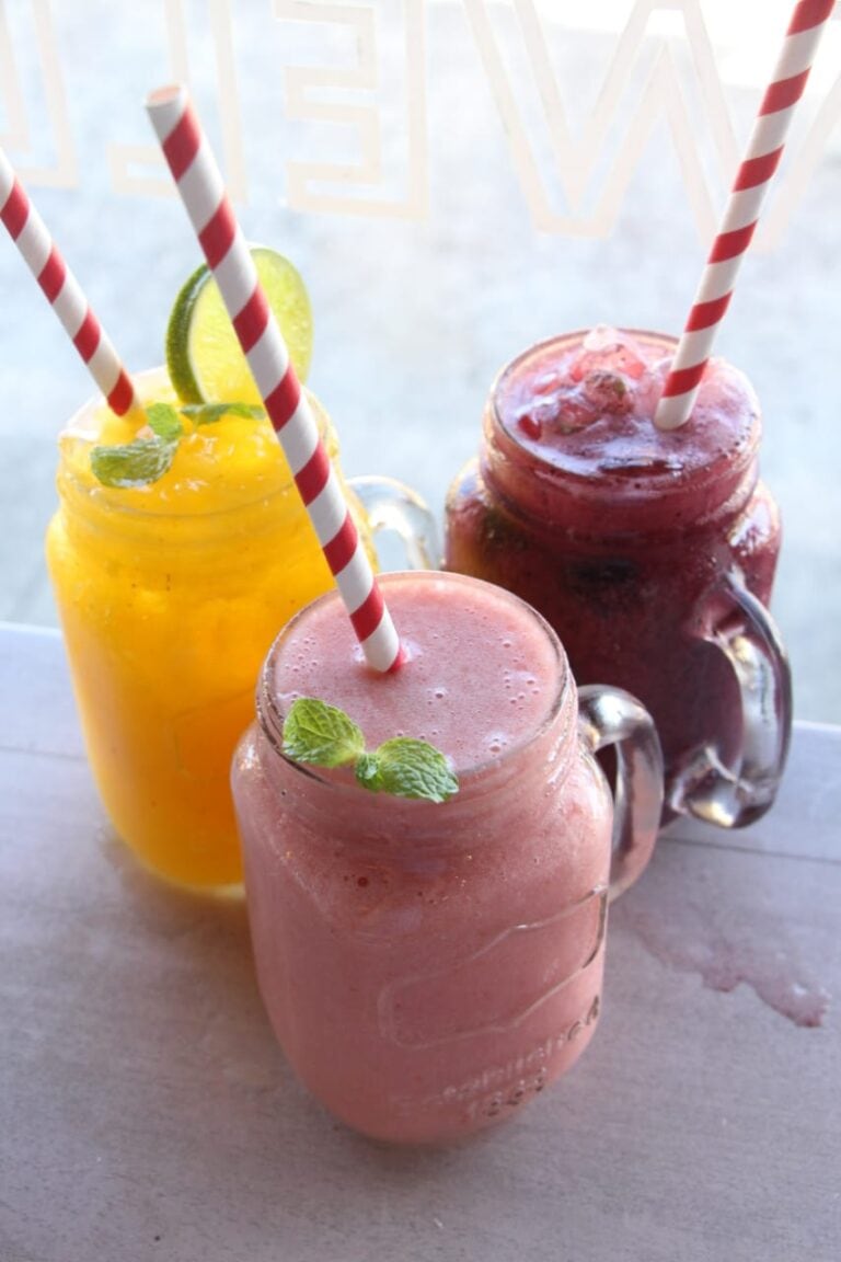Three colorful smoothies with striped paper straws and mint garnishes are displayed in mason jar glasses on a wooden surface, set against a light background with faint "Roatan" signage, representing a refreshing beverage option in Roatan, Honduras.
