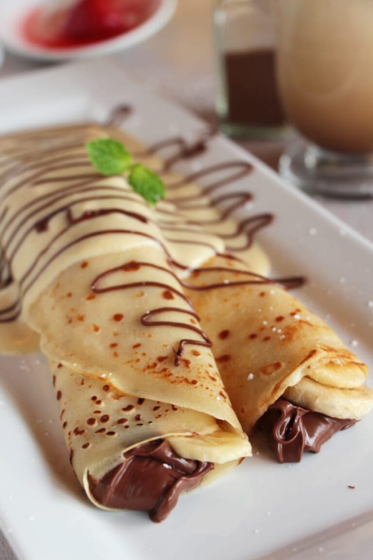 A plate of rolled crepes filled with chocolate, topped with chocolate drizzle and a sprig of mint is served on a white rectangular dish, with a blurred background showing a drink and other desserts at a restaurant on Roatan in Honduras.