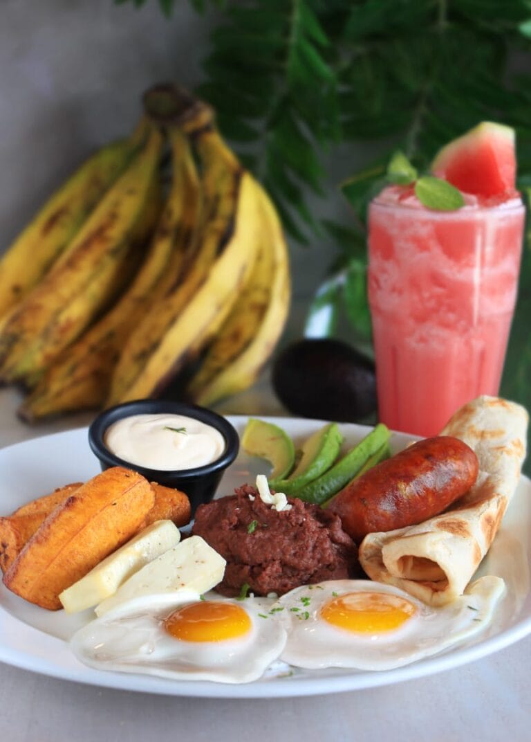 A traditional Honduran desayuno with fried eggs, refried beans, plantains, cheese, avocado, a tamale, and sausage is served on a white plate with a side of banana bunch and a pink fruit smoothie garnished with mint in the background, set against a tropical greenery backdrop in Roatan, Honduras.