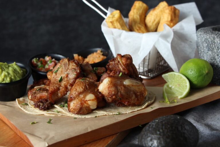 A plate of roasted chicken wings served with guacamole, salsa, and hot sauce, accompanied by fried plantains, lime wedges, and a basket of French fries on a wooden serving board in Roatan, Honduras.