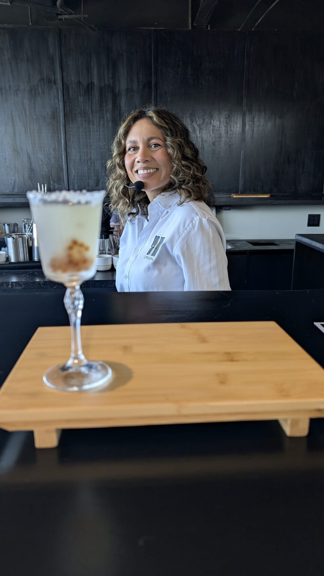 A woman with curly hair wearing a white shirt and headset, smiling behind a black counter in a bar or restaurant on Roatan, Honduras with black walls in the background.