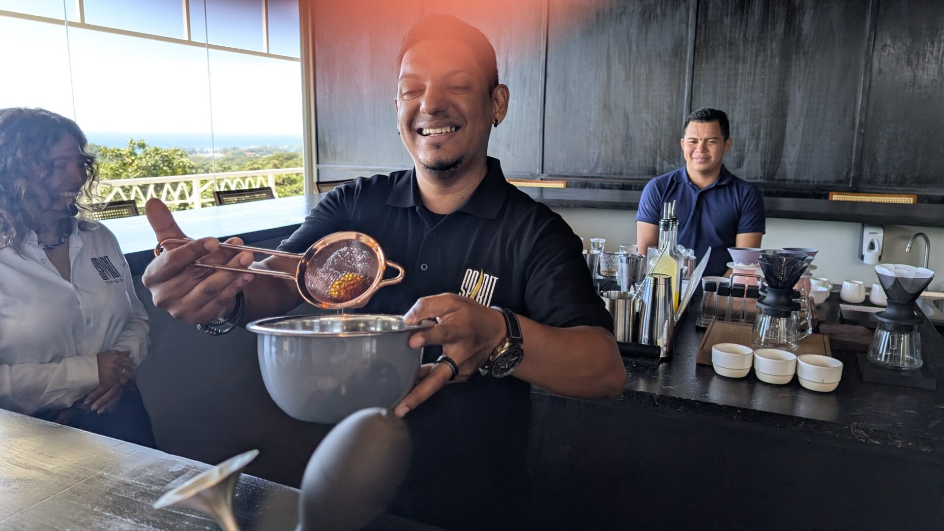 A smiling man in a black shirt holds a metal strainer over a mixing bowl while preparing drinks at a bar inside a restaurant on Roatan, Bay Islands, Honduras, with large windows showing a tropical landscape outside.