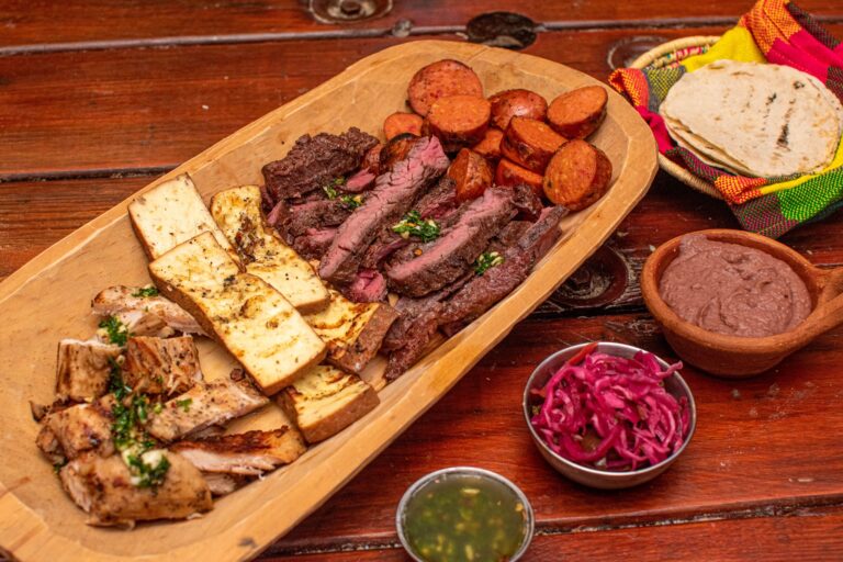 A wooden platter displays grilled steak, sausages, grilled cheese, and sliced chicken, accompanied by small bowls of sauce and pickled vegetables on a wooden table in Roatan, Honduras.