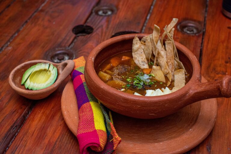 A bowl of traditional Honduran beef stew garnished with chopped herbs and accompanied by crispy totopos and sliced avocado sits on a colorful woven cloth on a wooden table in Roatan, Honduras.