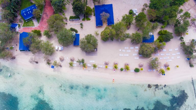 An aerial view of a beachside resort in Roatan, Honduras, featuring cabanas with blue roofs, oceanfront lounge chairs, and palm trees lining the sandy shoreline.