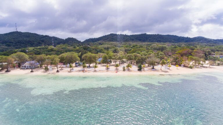 A beach with white sand, greenish turquoise water, and tropical trees lining the shore on Roatan in Honduras, with hills and cloudy sky in the background.
